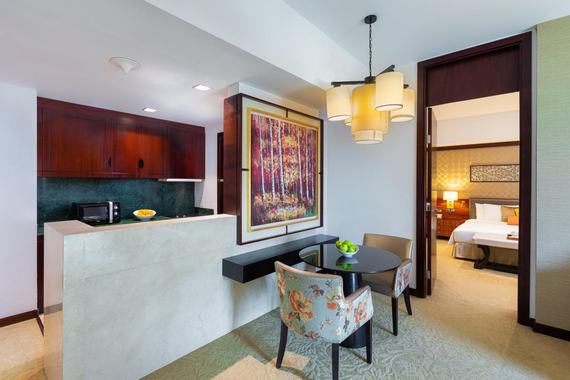 Dusit Thani דובאי מראה חיצוני תמונה The photo shows a well-furnished hotel room or suite. On one side, there is a small kitchenette with dark wooden cabinets and a countertop. A microwave and fruits are visible on the counter. 

Adjacent to the kitchenette is a dining area featuring a 