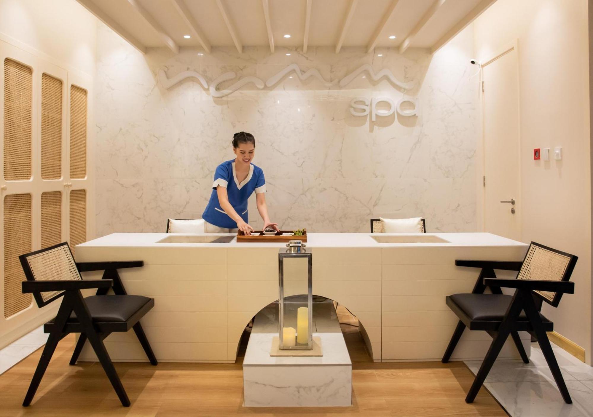 Dusit Thani דובאי מראה חיצוני תמונה The photo depicts a serene spa reception area. In the center, a staff member is seen preparing for services at a modern reception desk adorned with a marble surface. The background features a sleek, light-colored design with the word "spa" elegantly 